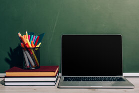 laptop with blank screen and pen holder with stationery on stack of books near green chalkboard